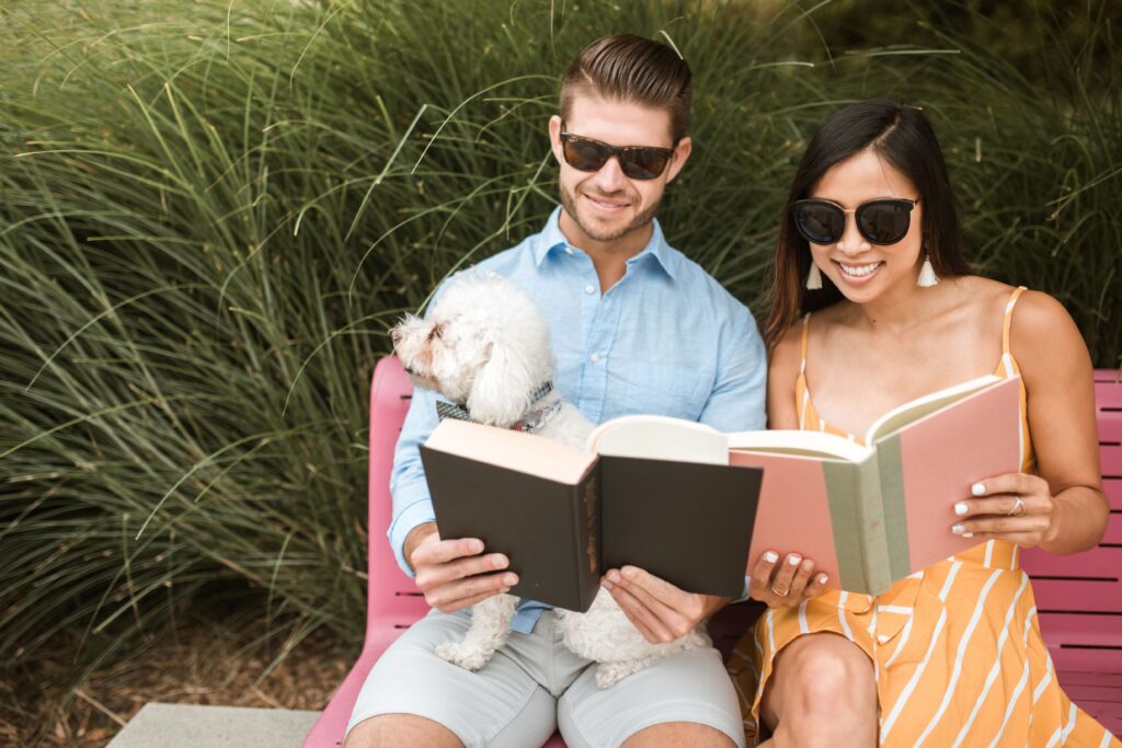 Happy couple reading books together with their dog on a vibrant pink bench in a sunny park.