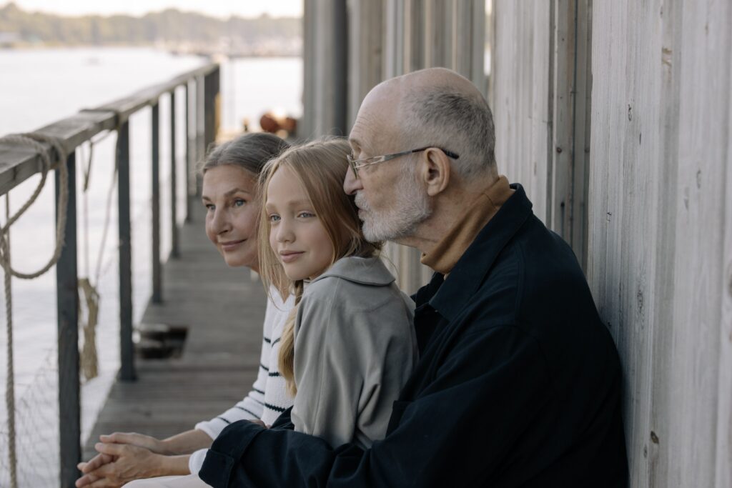 A loving family moment with grandparents and granddaughter sitting by the waterfront, outdoors.