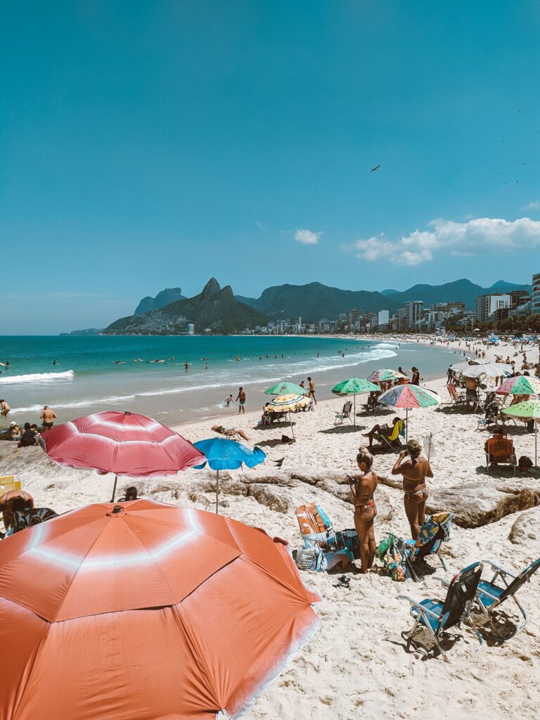 Anonymous tourists sunbathing on sandy shore with parasols against sea and mount under cloudy blue sky in sunlight