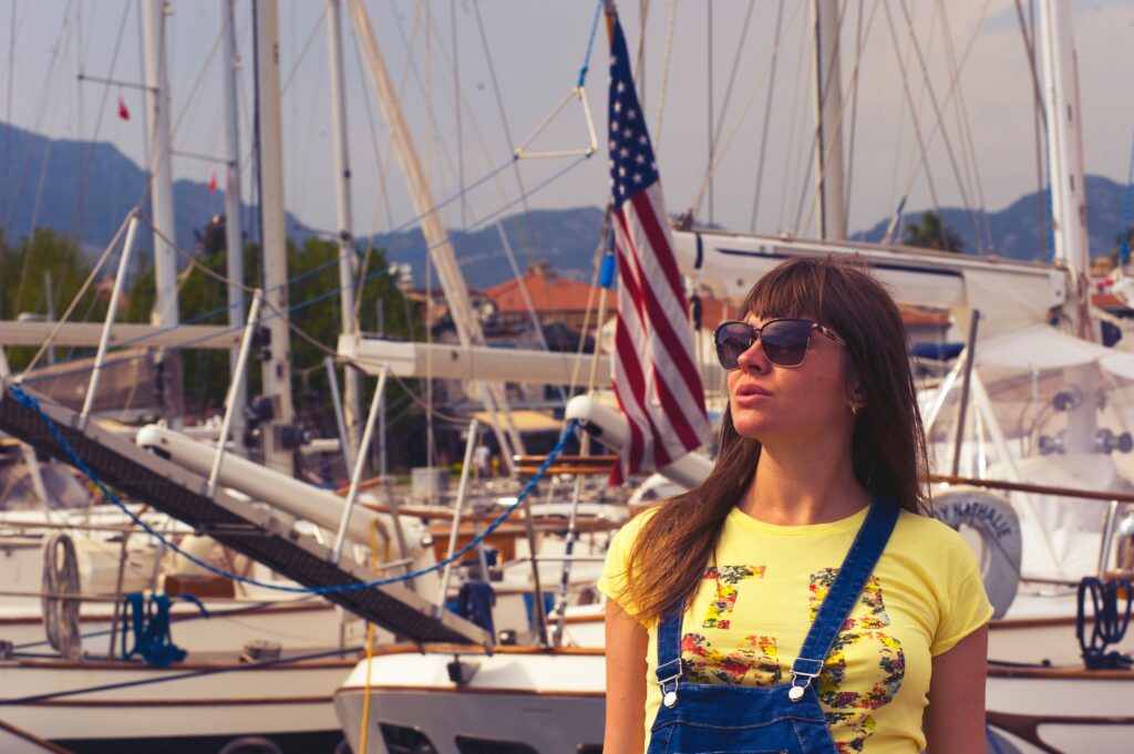 A woman in sunglasses stands at the marina in Marmaris, Turkey, with sailboats and an American flag in the background.