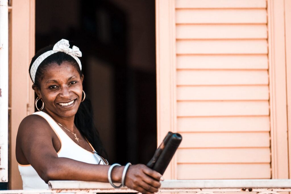 Smiling woman with a headband leaning out of a shuttered window on a sunny day.