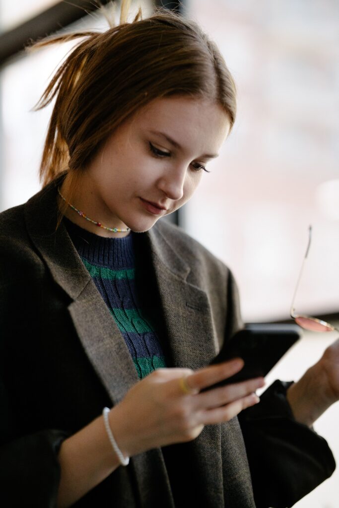 A young woman in a blazer using her smartphone indoors, holding eyeglasses.