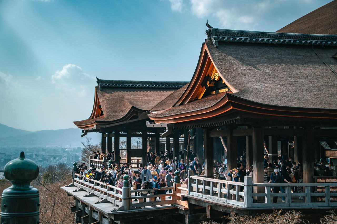 Tourists gather on the sunlit terraces of Kiyomizu-dera Temple in Kyoto, Japan, under a clear blue sky.