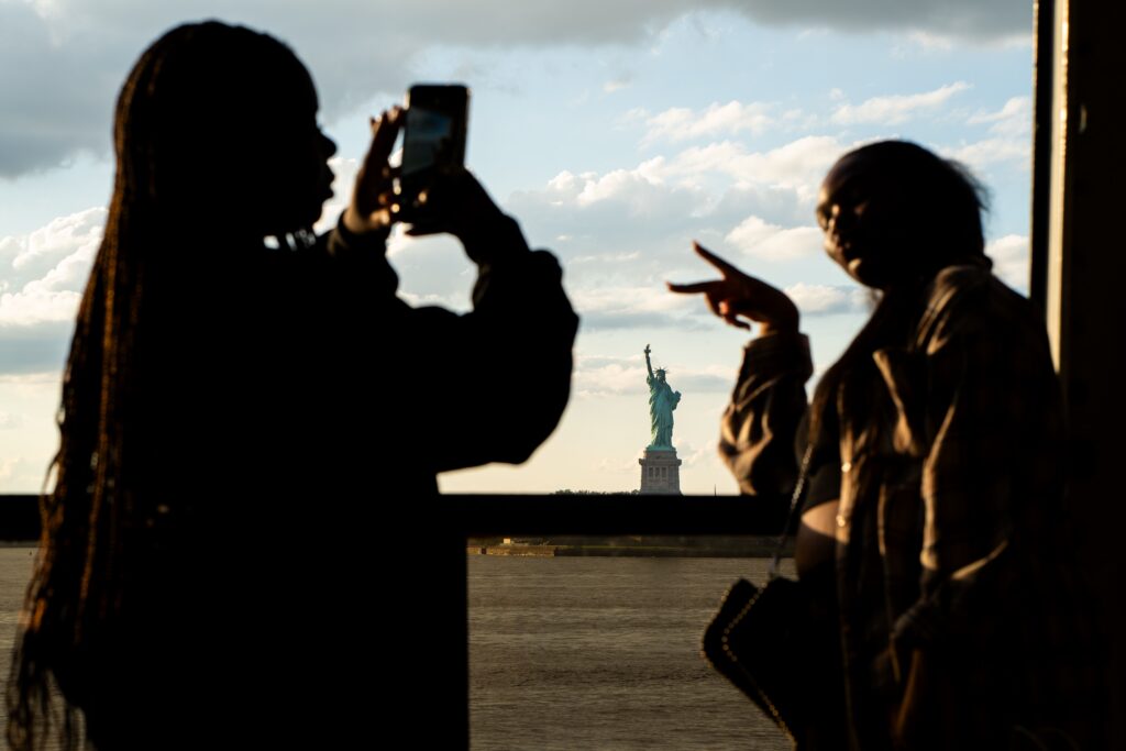 Silhouettes of tourists capturing the iconic Statue of Liberty at sunset in New York City.