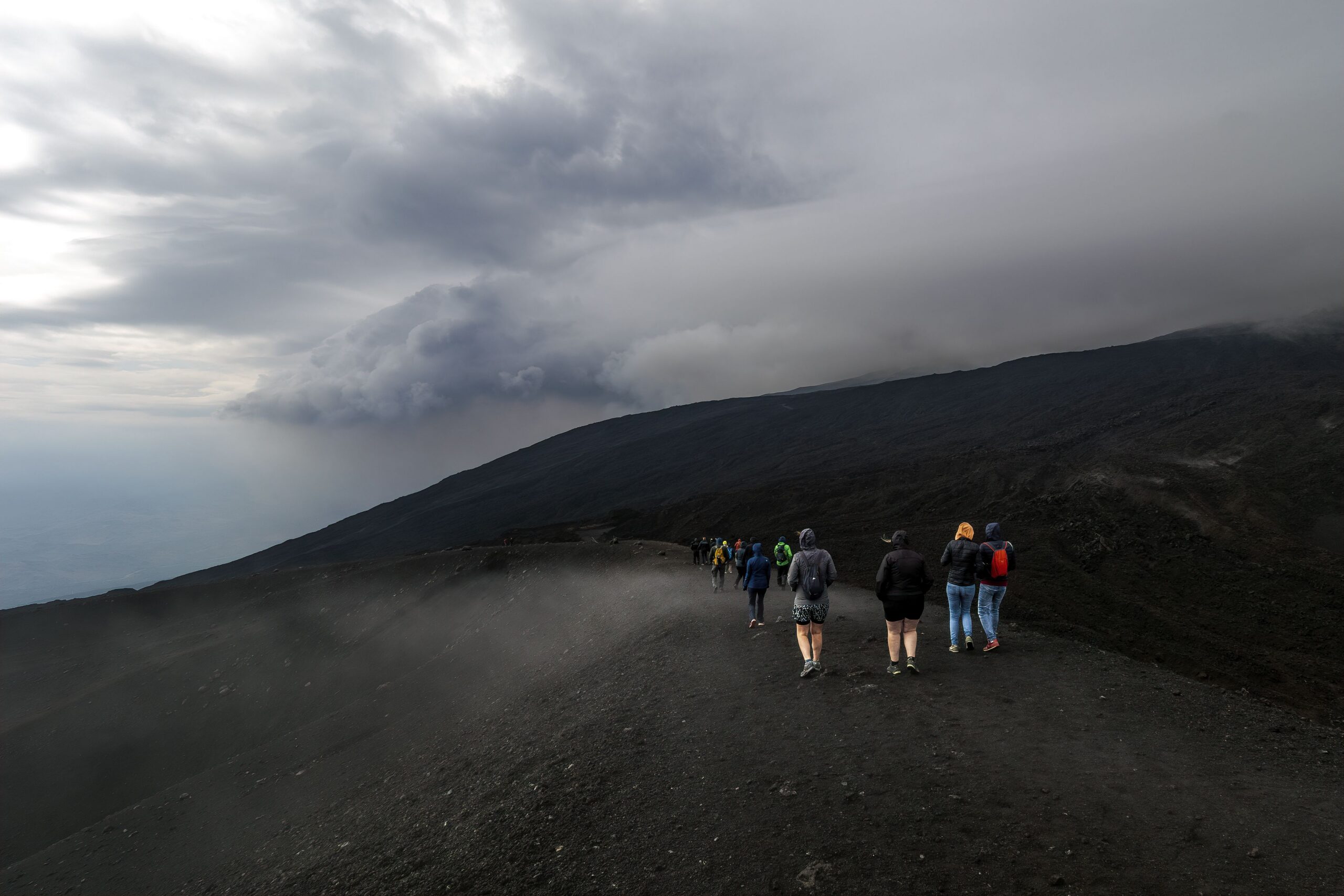Group of people hiking on a volcanic landscape under a dramatic cloudy sky, creating a moody atmosphere.