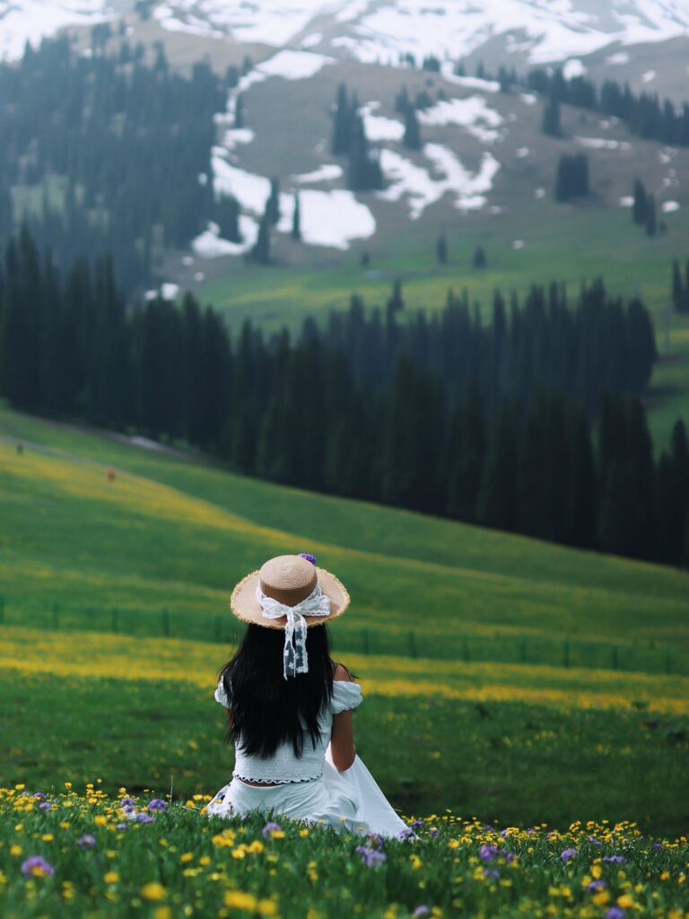 Woman wearing a straw hat sits in a meadow, overlooking scenic mountains and forests in Xinjiang, China.