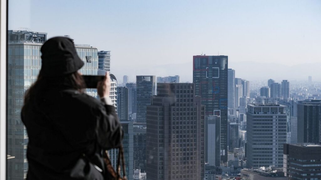 A person photographs the stunning Osaka skyline from a high-rise building window, showcasing urban architecture.