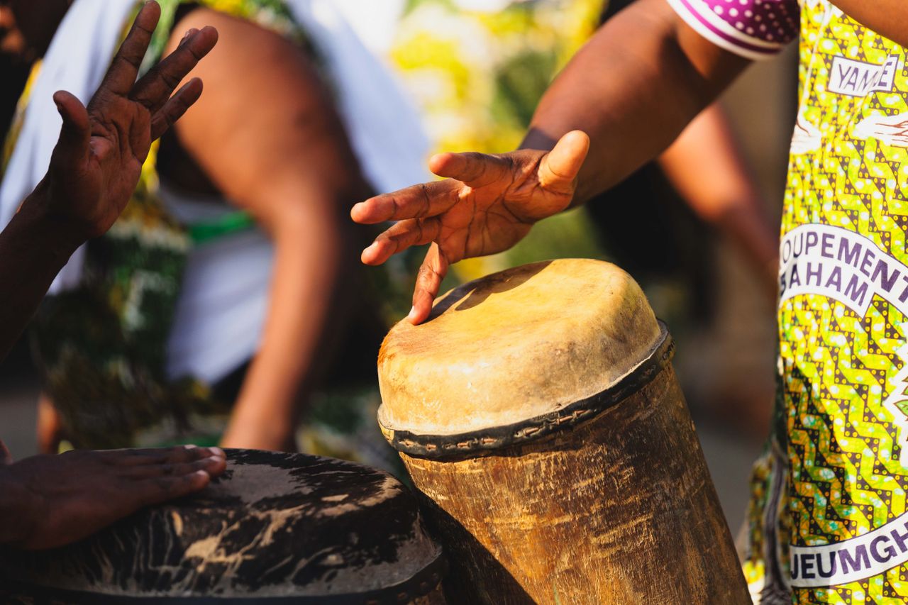 Hand drumming on traditional African drums during an outdoor ceremony, colorful attire, cultural expression.