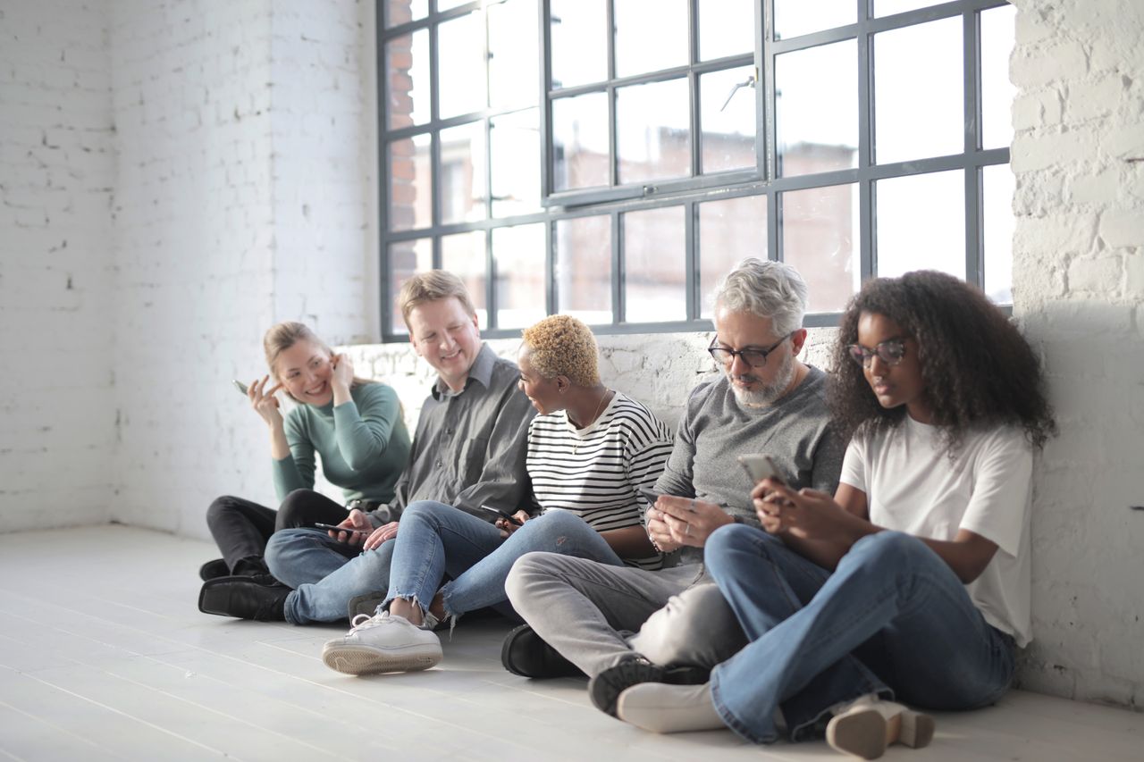 Cheerful multiethnic coworkers of different ages surfing internet on cellphones while sitting with crossed legs near brick wall and fenced window during daytime