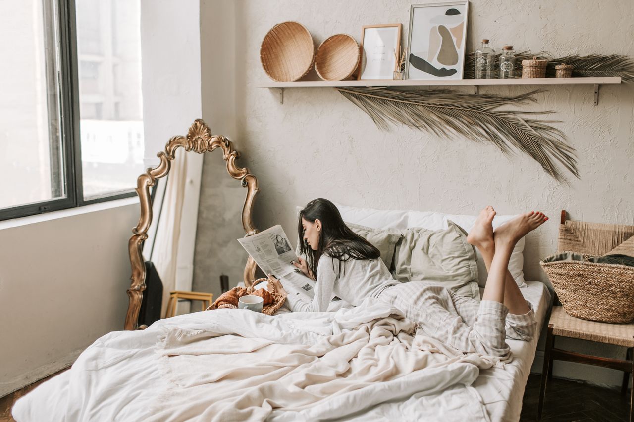 Woman reading a newspaper in a cozy bedroom setting with a decorative mirror and natural lighting.