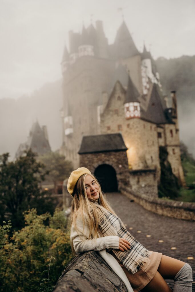 A woman poses in front of the fog-covered Eltz Castle in Germany, creating a dreamy travel scene.