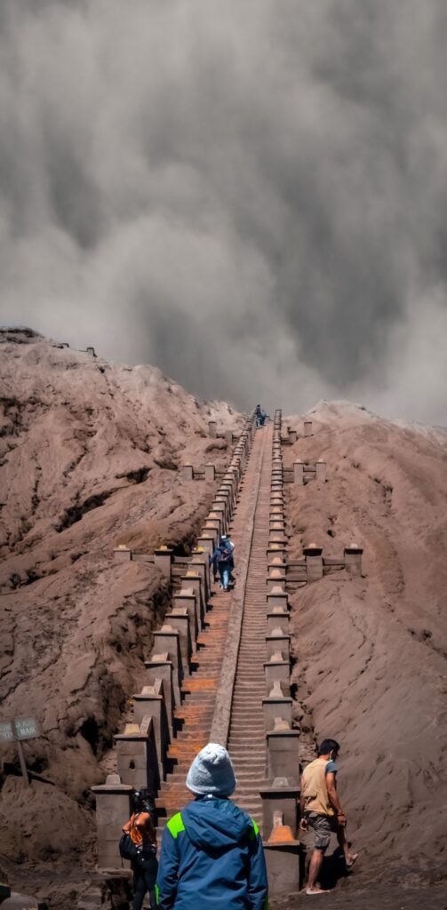 Hikers ascend a dramatic staircase, surrounded by volcanic ash, under a cloudy sky.