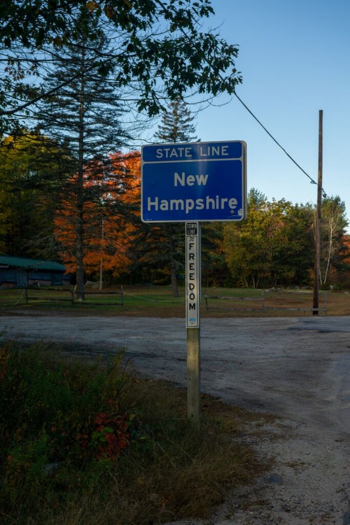 Blue New Hampshire state line sign with autumn foliage at roadside.