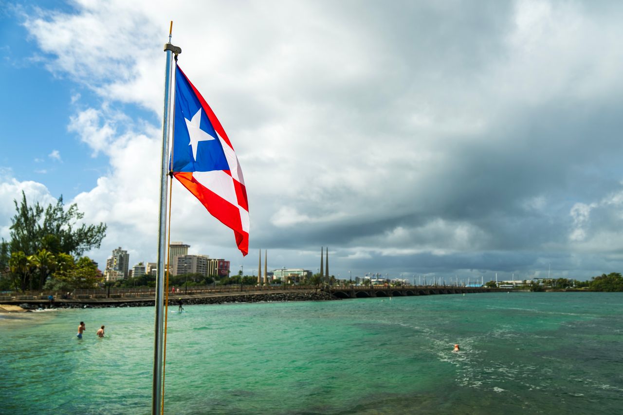 Puerto Rican flag on a flagpole by the sea with city skyline in the background.
