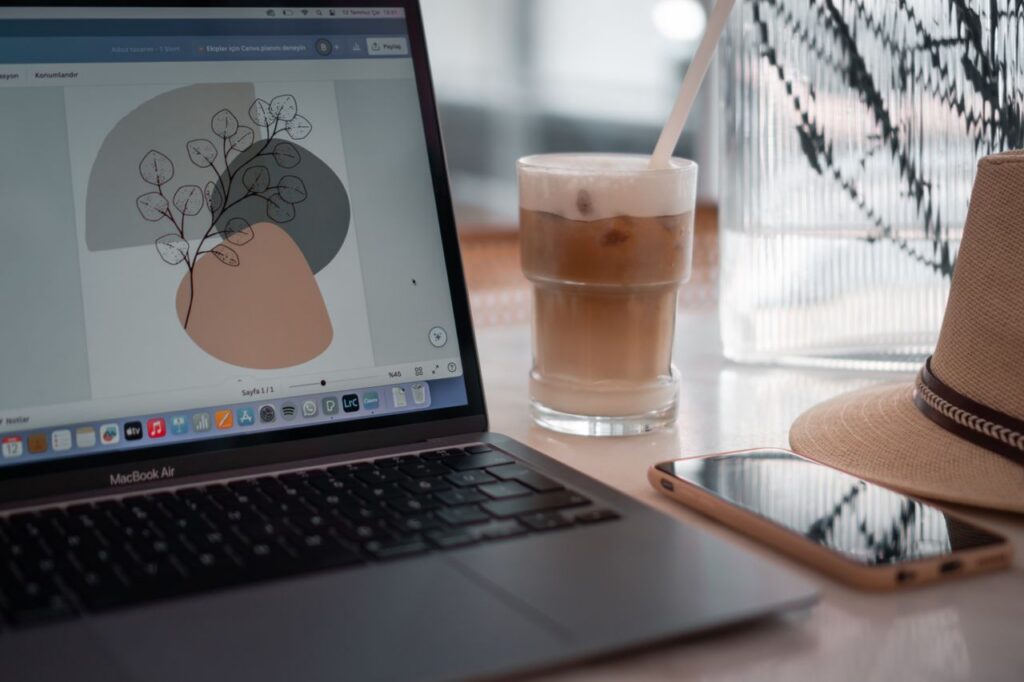 A modern workspace featuring a laptop, iced coffee, smartphone, and hat on a table.
