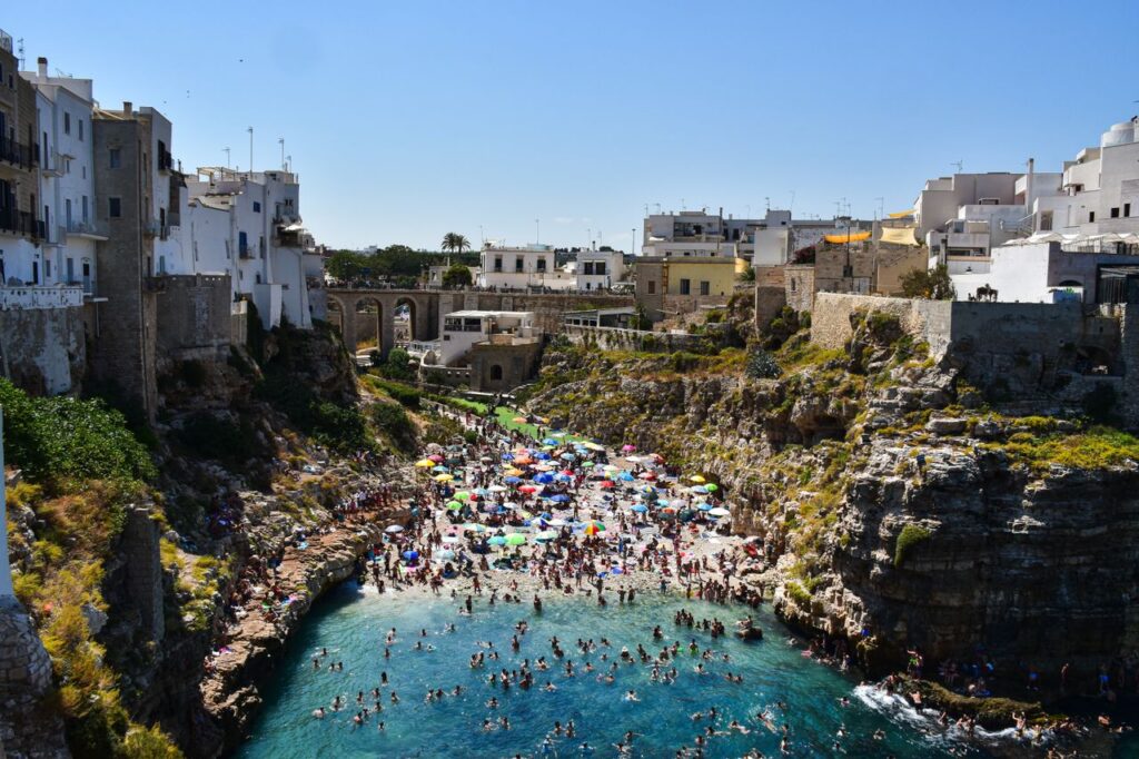 Vibrant summer scene of a crowded beach surrounded by cliffs in Polignano a Mare, Italy.