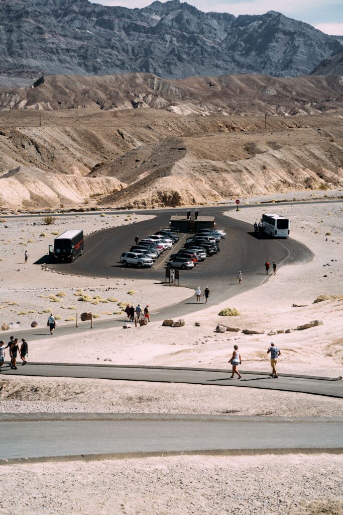 Tourists explore a desert parking area with cars and buses, set against mountain backdrop.